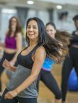 A multi-ethnic group of adult women are dancing in a fitness studio. They are wearing athletic clothes. An Ethnic woman is smiling while dancing in the foreground.