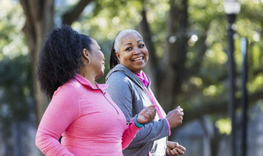 Two women exercising together in the city, jogging or power walking, laughing and conversing. Buildings and trees are out of focus in the background. The one in pink is in her 60s and her friend is in her 50s.