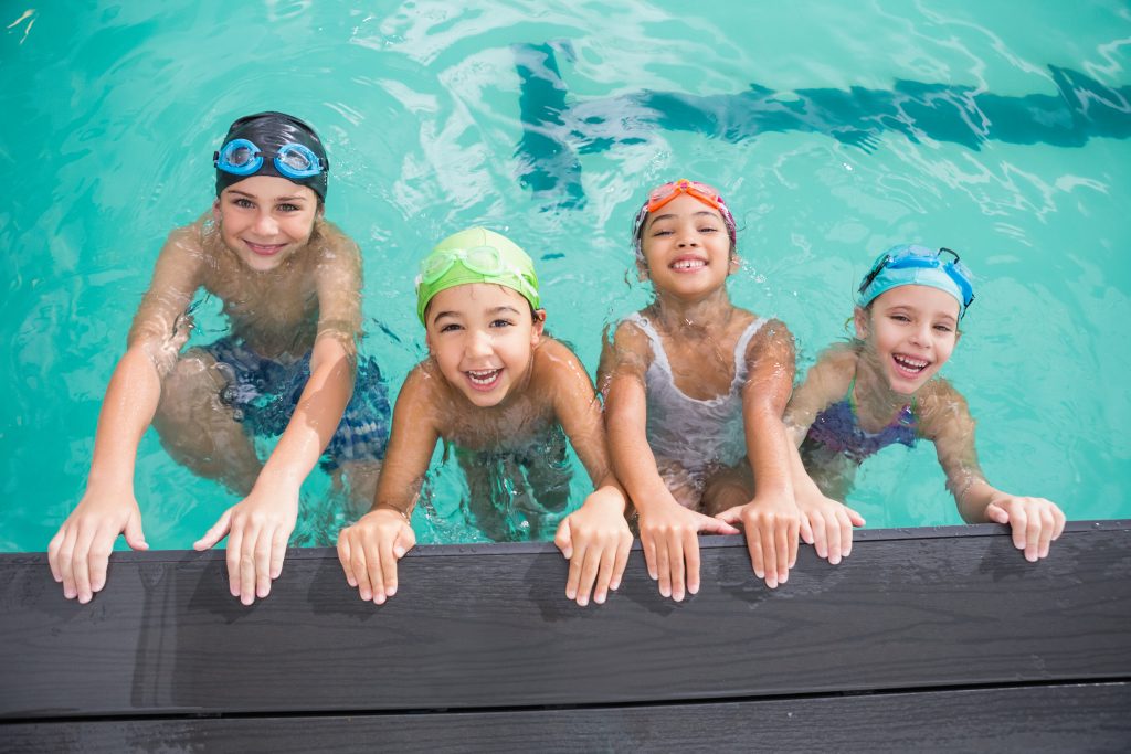 A group of four young people shown in a swimming pool. They are holding on to the side of the pool smiling as they wait to start their swimming lesson