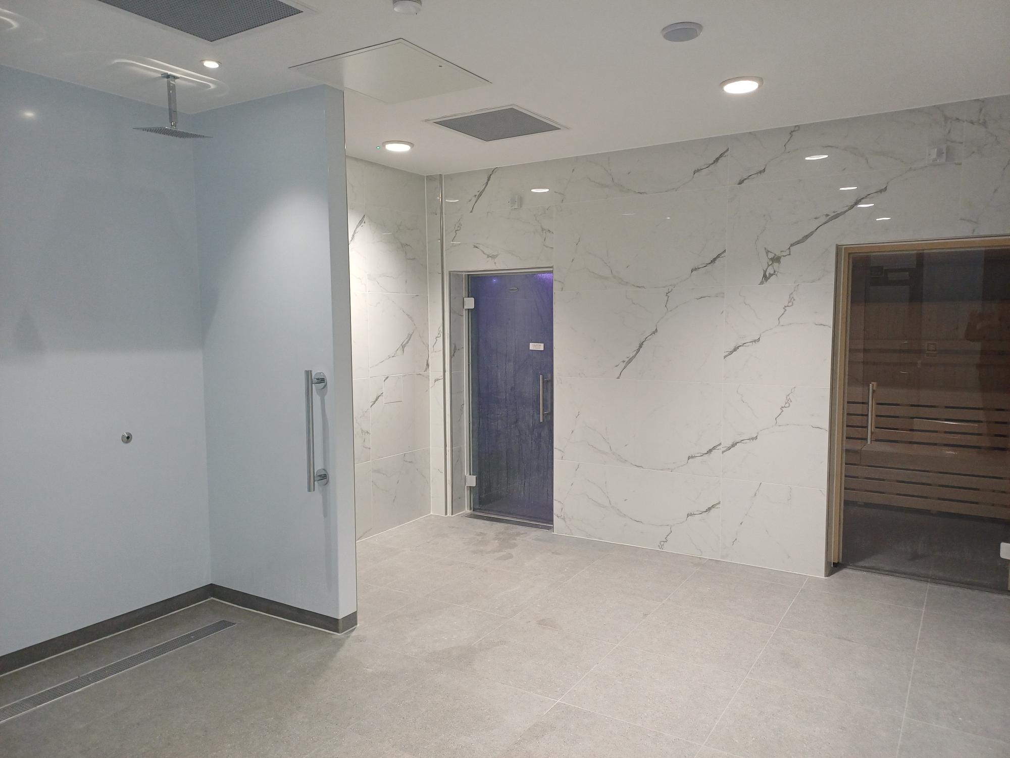 New waterfall shower cubicles, a marble style wall with two doors. The left door leading to the steam room and the right door leading to the sauna.