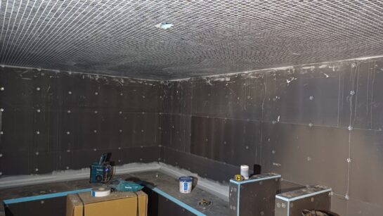 The thermal suite area at Rochdale Leisure Centre under construction, showing the ceiling covered in a grid of small white tiles. The lower half of the walls are dark panels, with visible fasteners, while the upper sections are bare concrete. There are various construction materials and tools on the floor. The sauna bench is partly constructed.