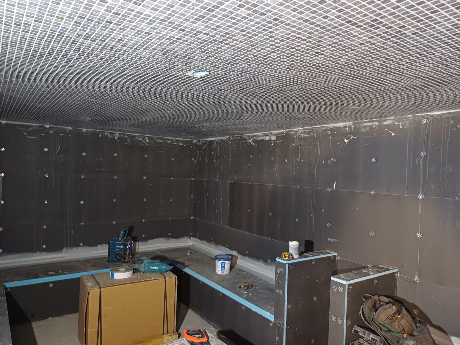 The thermal suite area at Rochdale Leisure Centre under construction, showing the ceiling covered in a grid of small white tiles. The lower half of the walls are dark panels, with visible fasteners, while the upper sections are bare concrete. There are various construction materials and tools on the floor. The sauna bench is partly constructed.