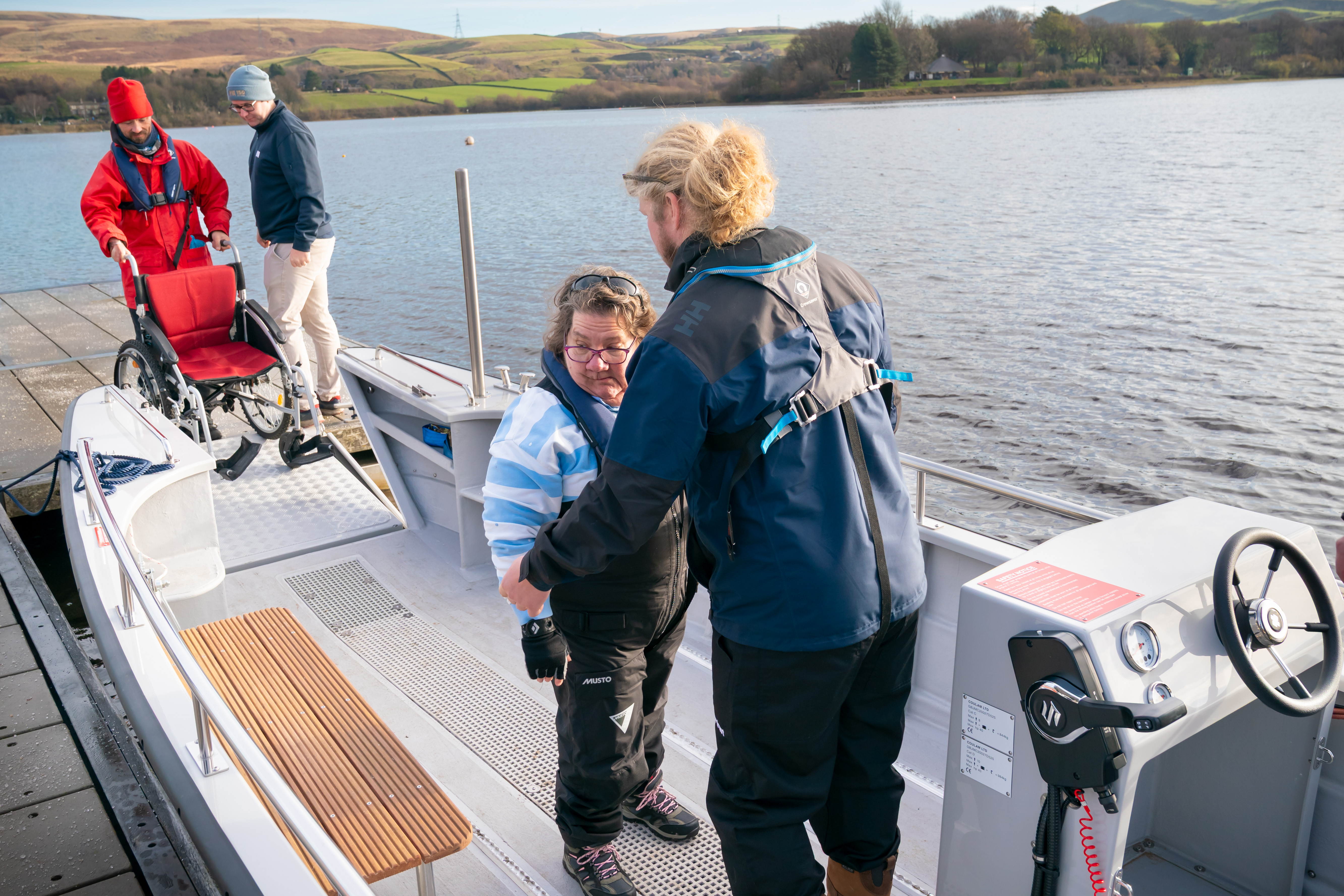 Two people stand inside the wheelchair-accessible Wheelyboat at Hollingworth Lake, with one assisting the other near the seating area. On the dock, another person is preparing a red wheelchair for boarding. The boat’s steering console is visible on the right, and the lake with Pennine hills forms the scenic background.