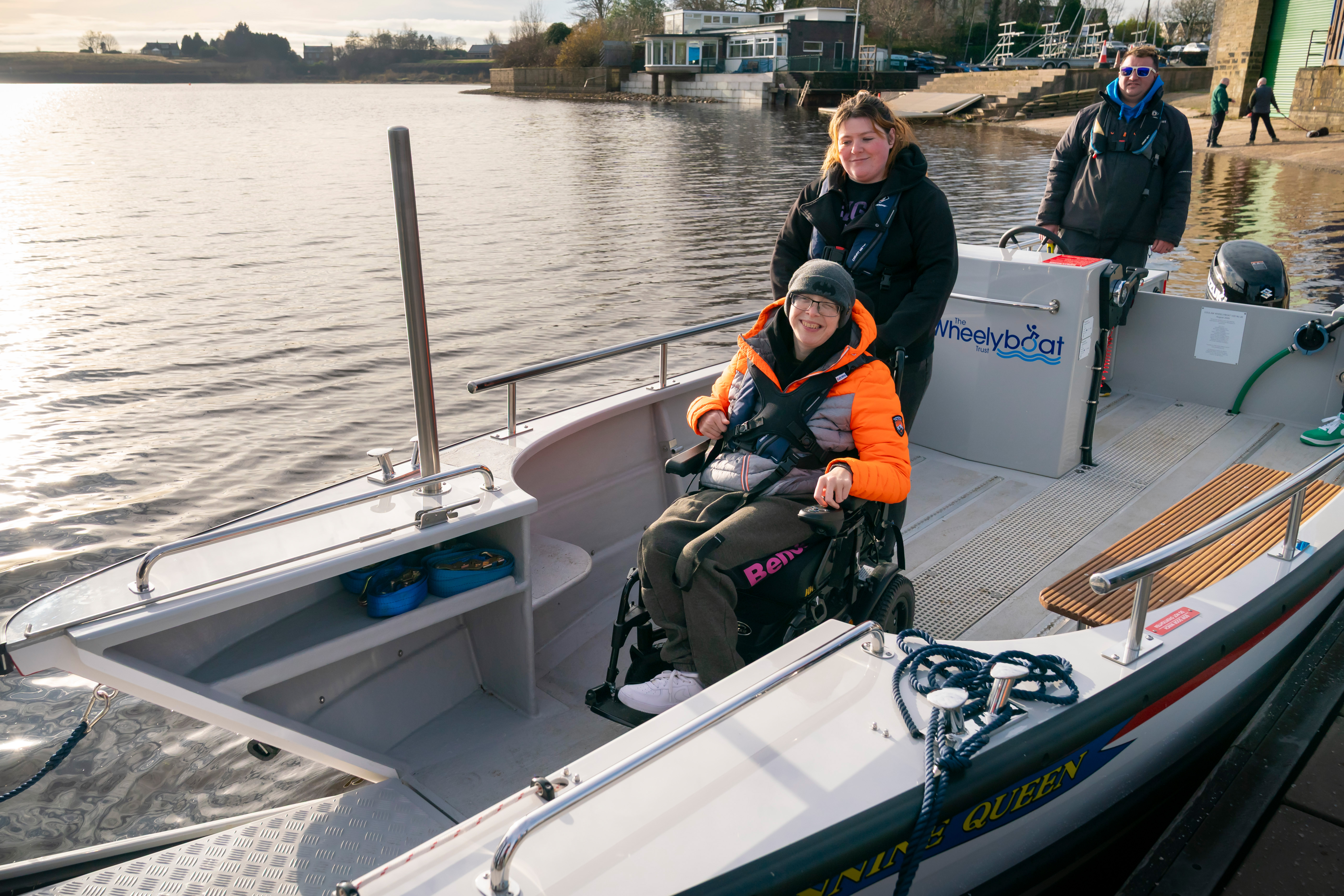 A person seated in a wheelchair on the newly launched Wheelyboat named ‘Pennine Queen’ at Hollingworth Lake, wearing a bright orange jacket and secured with a safety belt. Two other individuals stand nearby on the boat, which is docked close to the shore with buildings and water in the background.