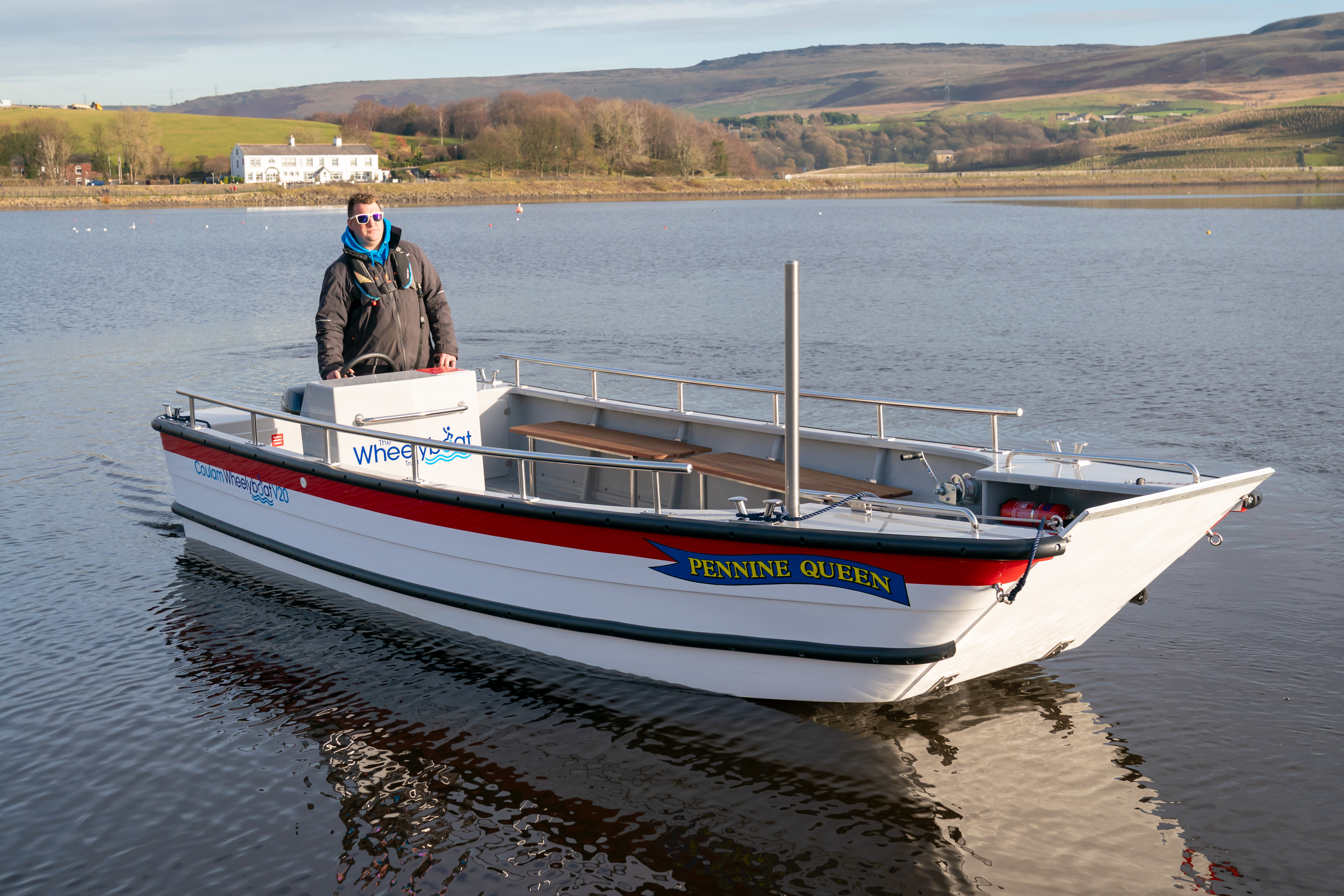 The newly launched wheelchair-accessible Wheelyboat named ‘Pennine Queen’ floating on Hollingworth Lake. A member of the Hollingworth Lake team who will drive the boat stands on the boat near the steering console, and the surrounding landscape features calm water, grassy hills, and a white building in the background.