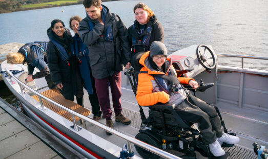 A group of passengers on the newly launched wheelchair-accessible Wheelyboat at Hollingworth Lake. One passenger is seated in a powered wheelchair near the steering console, while others stand on the boat wearing life jackets. The boat is docked beside the lake with the scenic Pennine hills in the background.