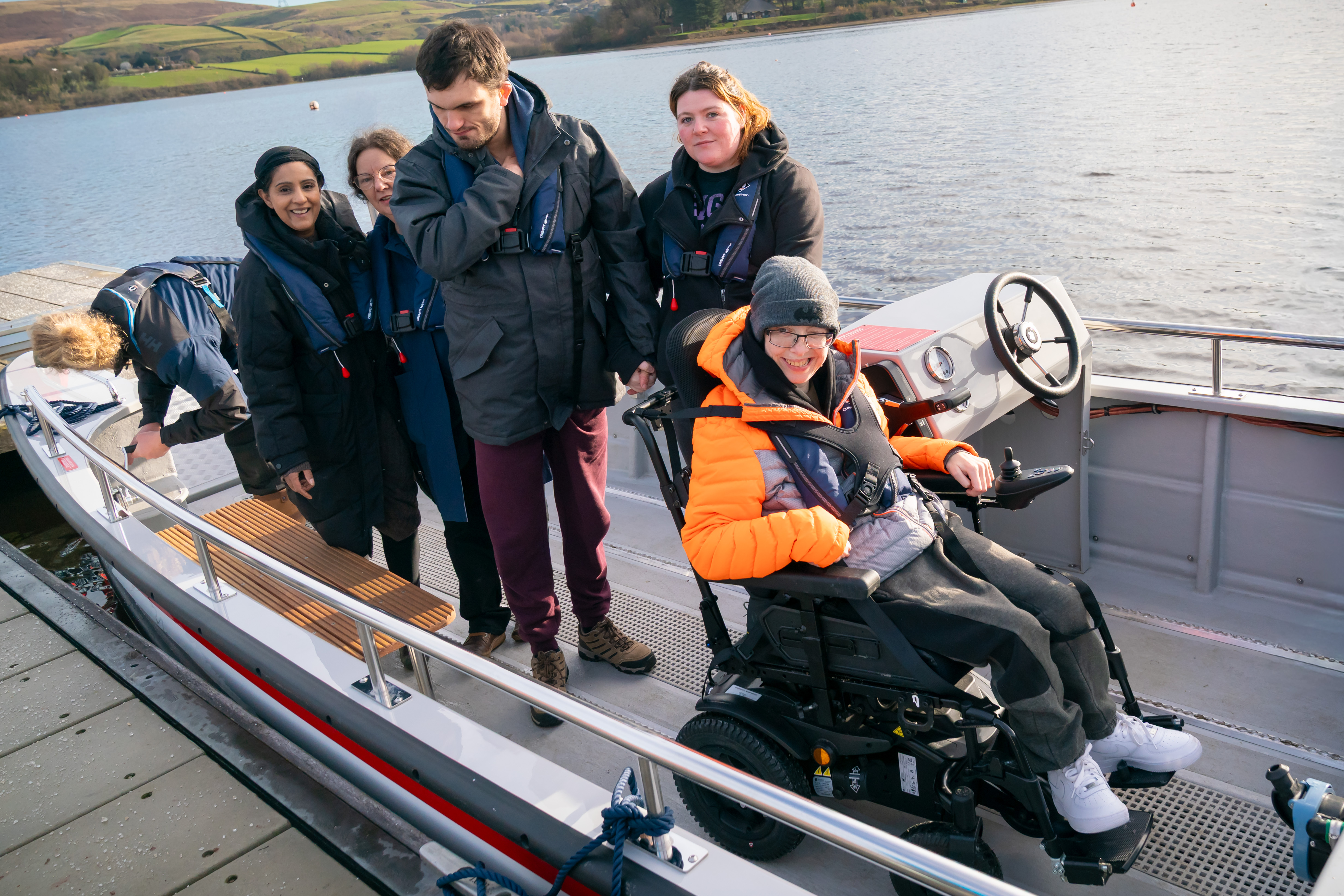 A group of passengers on the newly launched wheelchair-accessible Wheelyboat at Hollingworth Lake. One passenger is seated in a powered wheelchair near the steering console, while others stand on the boat wearing life jackets. The boat is docked beside the lake with the scenic Pennine hills in the background.