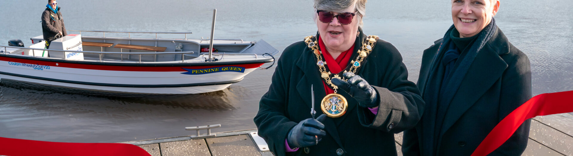 Rochdale Civic Mayor cuts a red ribbon to officially launch the new wheelchair-accessible Wheelyboat named ‘Pennine Queen’ at Hollingworth Lake. Standing beside the Mayor is Your Trust CEO Estelle Rowe. The boat is visible on the water in the background, with the scenic Pennine hills beyond.