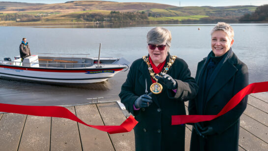 Rochdale Civic Mayor cuts a red ribbon to officially launch the new wheelchair-accessible Wheelyboat named ‘Pennine Queen’ at Hollingworth Lake. Standing beside the Mayor is Your Trust CEO Estelle Rowe. The boat is visible on the water in the background, with the scenic Pennine hills beyond.