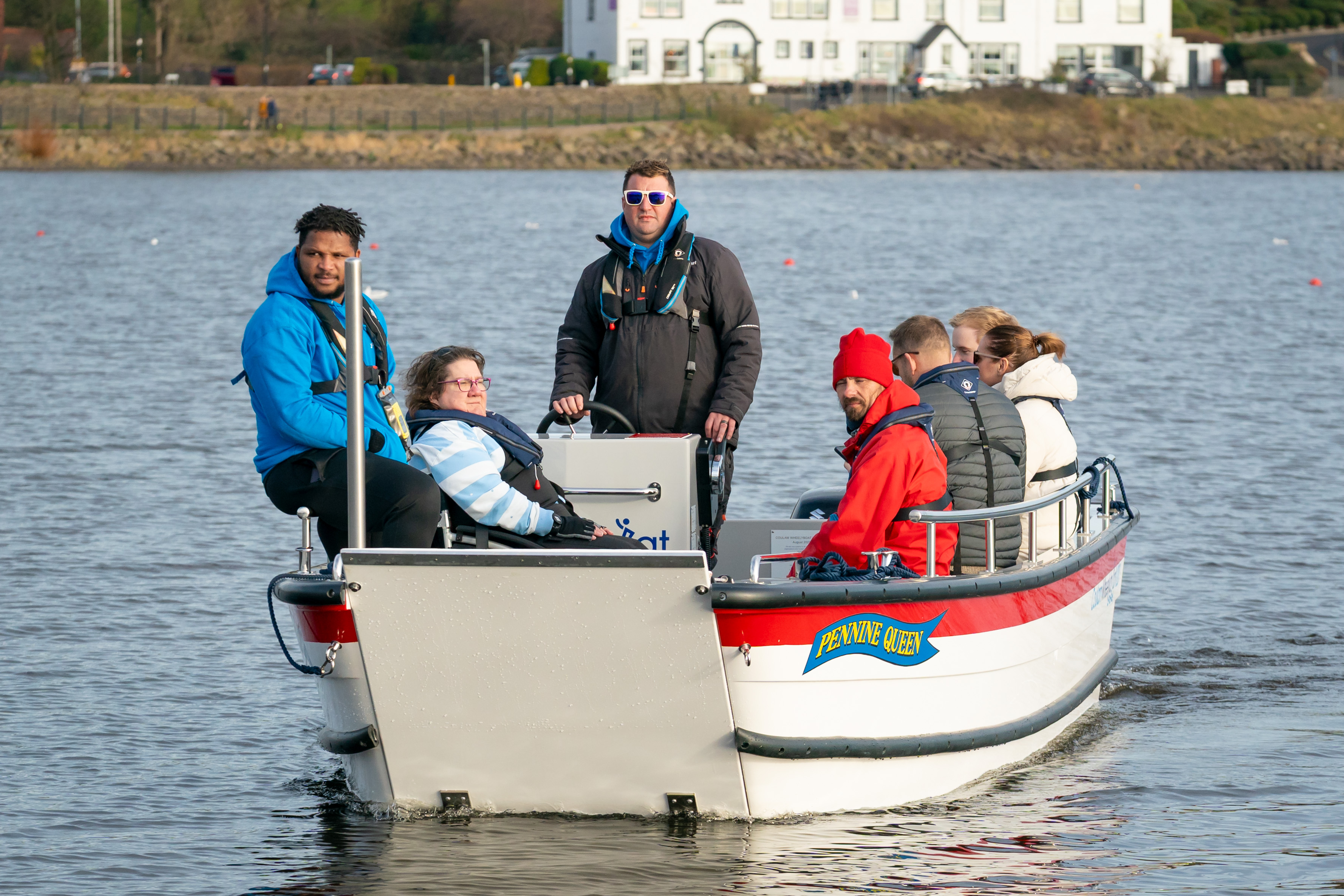 Participants enjoying a first ride on the newly launched wheelchair-accessible Wheelyboat named ‘Pennine Queen’ at Hollingworth Lake. The boat is on the water with several people seated and wearing life jackets, and a building is visible on the shoreline in the background.