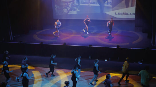 A group fitness class takes place at the Les Mills launch event at Middleton Arena. Three instructors perform choreography on a stage under colourful lighting, while a larger group of participants follow along on the floor in front of them. A large Les Mills-branded screen is projected behind the stage