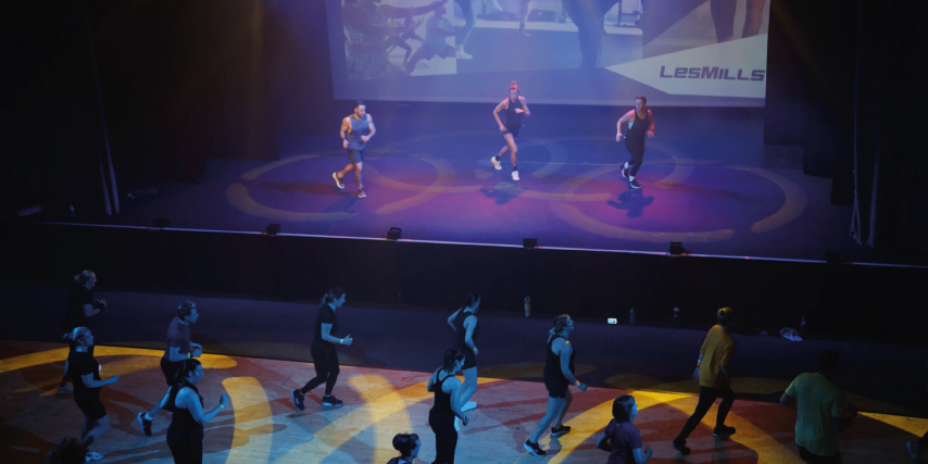 A group fitness class takes place at the Les Mills launch event at Middleton Arena. Three instructors perform choreography on a stage under colourful lighting, while a larger group of participants follow along on the floor in front of them. A large Les Mills-branded screen is projected behind the stage