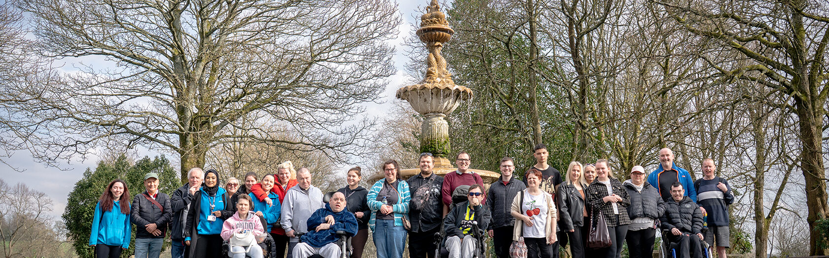 A large group of people taking part in a TfGM and Your Trust “Walks and Wheel” session are gathered on a wide path in a park. Some participants are standing, while others are using wheelchairs or mobility aids. The group is positioned in front of a tall, decorative stone fountain surrounded by leafless trees.