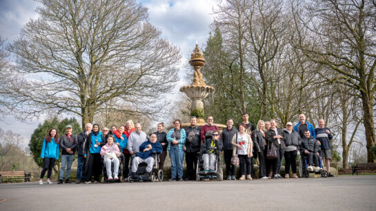A large group of people taking part in a TfGM and Your Trust “Walks and Wheel” session are gathered on a wide path in a park. Some participants are standing, while others are using wheelchairs or mobility aids. The group is positioned in front of a tall, decorative stone fountain surrounded by leafless trees.