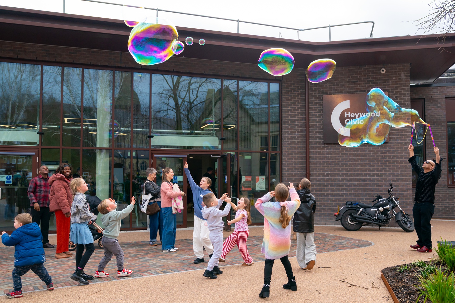 Families and children gather outside Heywood Civic during a community fun day for its reopening, with a bubble performer creating large, colourful soap bubbles in front of the building’s glass entrance and Heywood Civic sign.