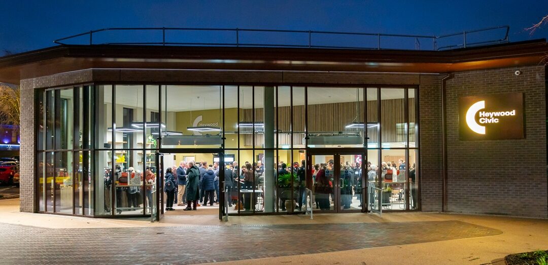 The exterior of the newly refurbished Heywood Civic at dusk, with a contemporary glass façade revealing people gathered inside, warm interior lighting, and an illuminated Heywood Civic sign on the brick frontage.