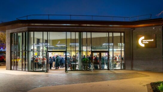 The exterior of the newly refurbished Heywood Civic at dusk, with a contemporary glass façade revealing people gathered inside, warm interior lighting, and an illuminated Heywood Civic sign on the brick frontage.