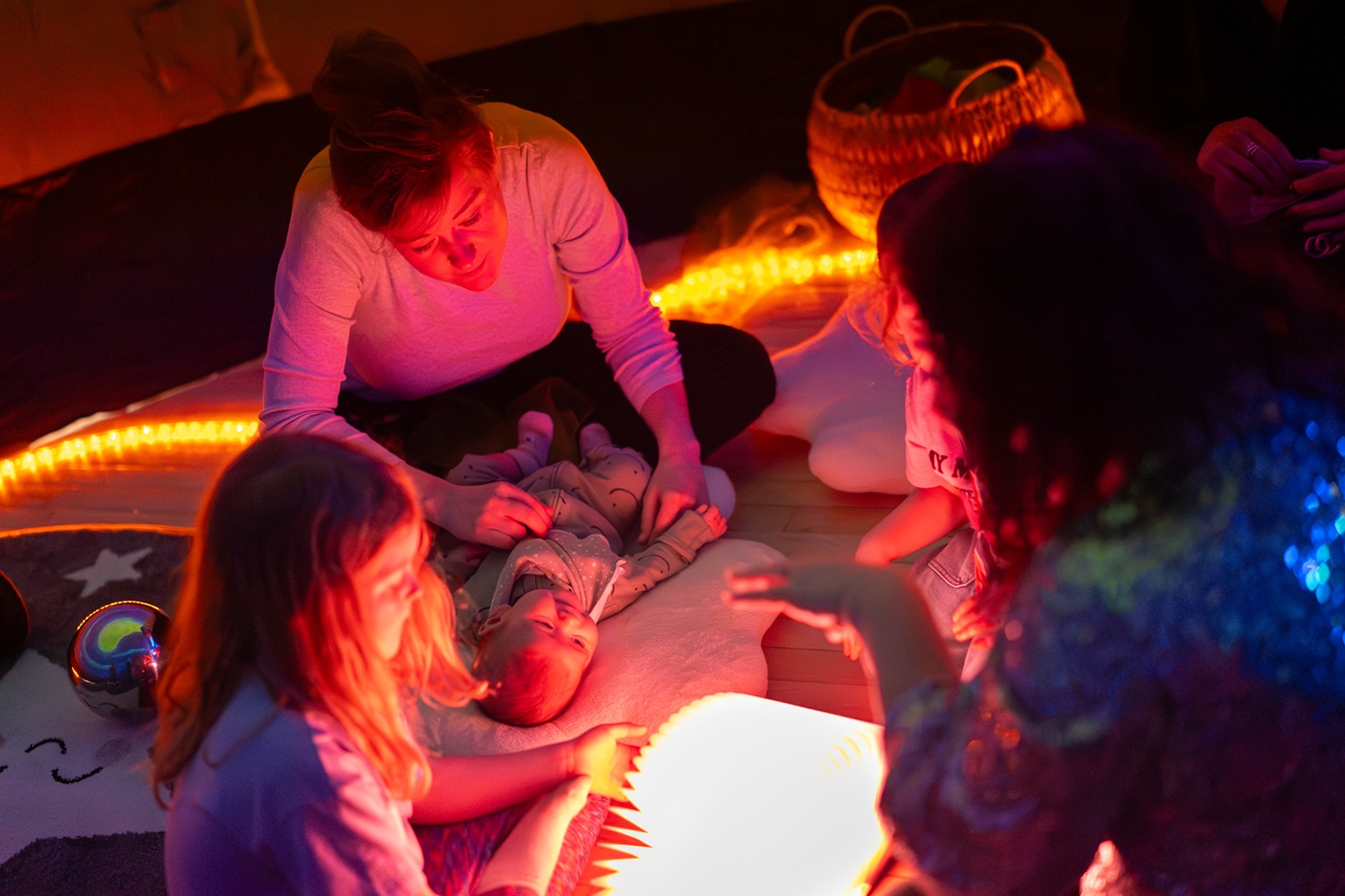 Indoor sensory activity at the community fun day for the Heywood Civic reopening, showing children and an adult seated on floor mats, interacting with soft lighting, cushions, and tactile play materials in a low-lit space.